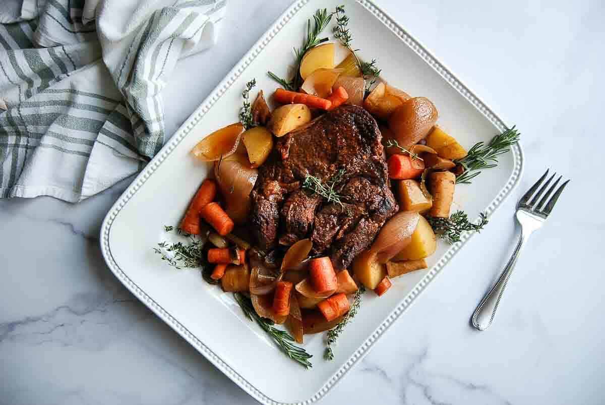 A roasted beef pot roast on a white platter, surrounded by cooked carrots, potatoes, onions, and garnished with fresh herbs.