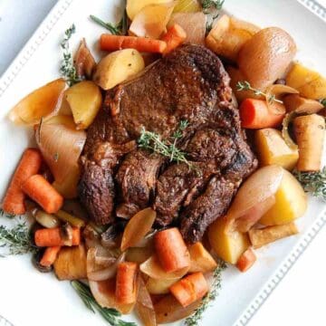 A cooked Dutch oven pot roast surrounded by carrots, potatoes, onions, and herbs is arranged on a white square plate.