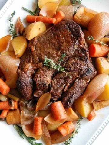 A cooked Dutch oven pot roast surrounded by carrots, potatoes, onions, and herbs is arranged on a white square plate.