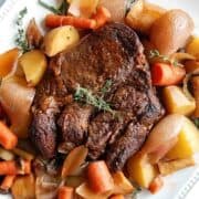 A cooked Dutch oven pot roast on a white plate, surrounded by roasted carrots, potatoes, onions, and fresh herbs such as rosemary and thyme.