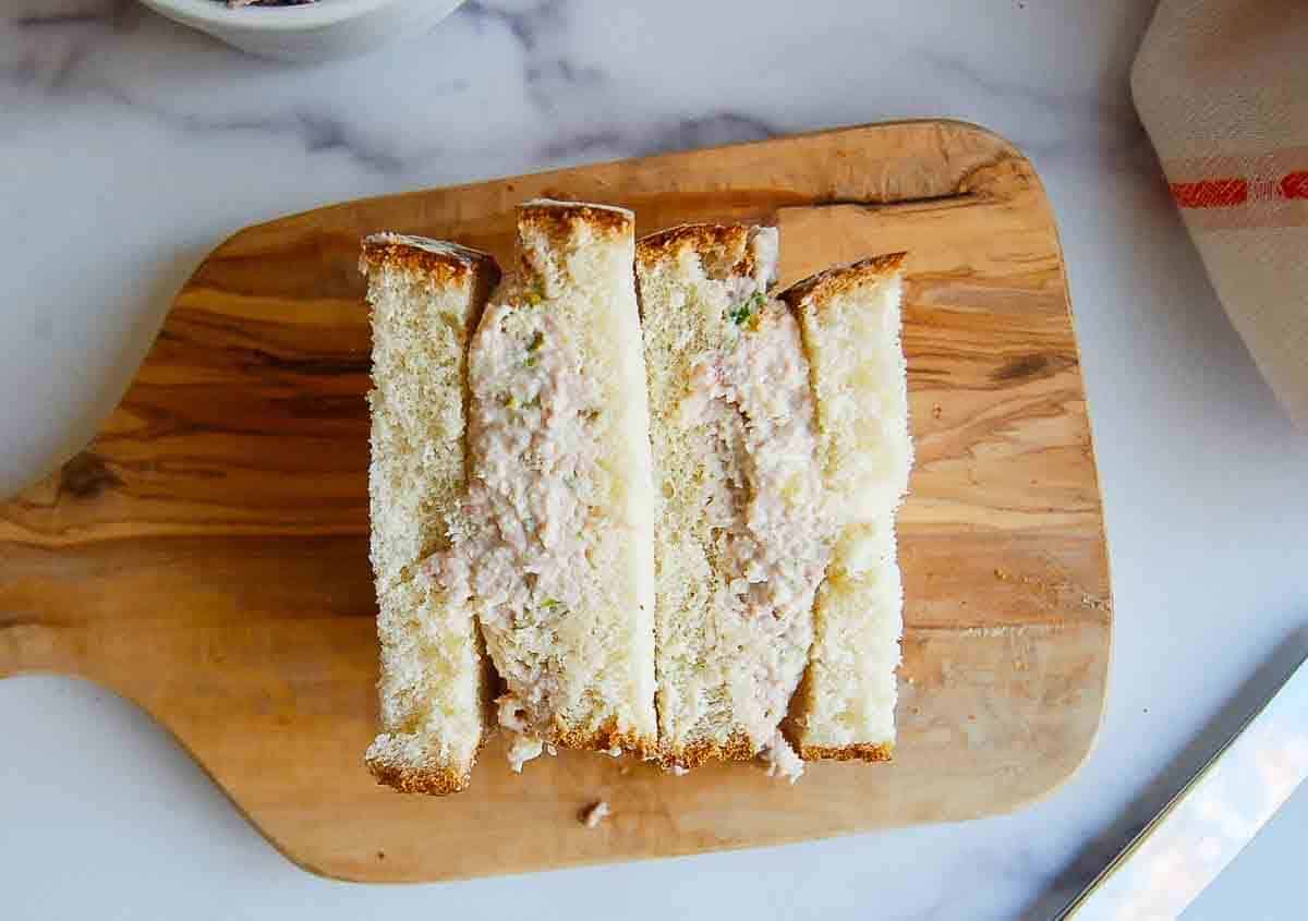 A wooden board holds three neatly sliced finger sandwiches filled with a creamy ham mixture, viewed from above on a light marble surface.