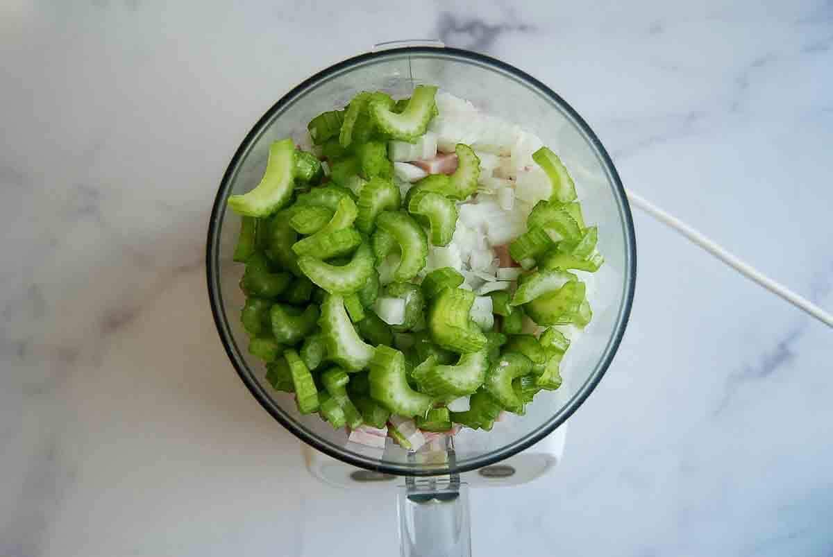 A food processor bowl filled with chopped celery, onions, and ham.