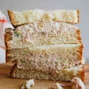 A close-up of three stacked ham salad sandwiches on white bread, with creamy ham filling visible and some filling spilling out onto a wooden surface.
