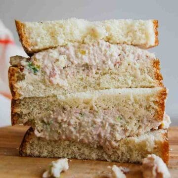 A close-up of three stacked ham salad sandwiches on white bread, with creamy ham filling visible and some filling spilling out onto a wooden surface.