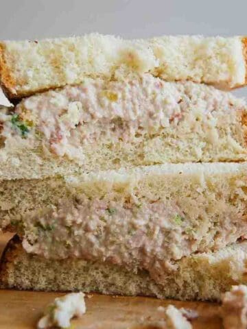 A close-up of three stacked ham salad sandwiches on white bread, with creamy ham filling visible and some filling spilling out onto a wooden surface.