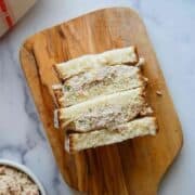 Three triangle-cut sandwiches filled with a creamy spread are stacked on a wooden cutting board. A knife and part of a cloth are visible nearby, along with a bowl of the same spread on a marble surface.