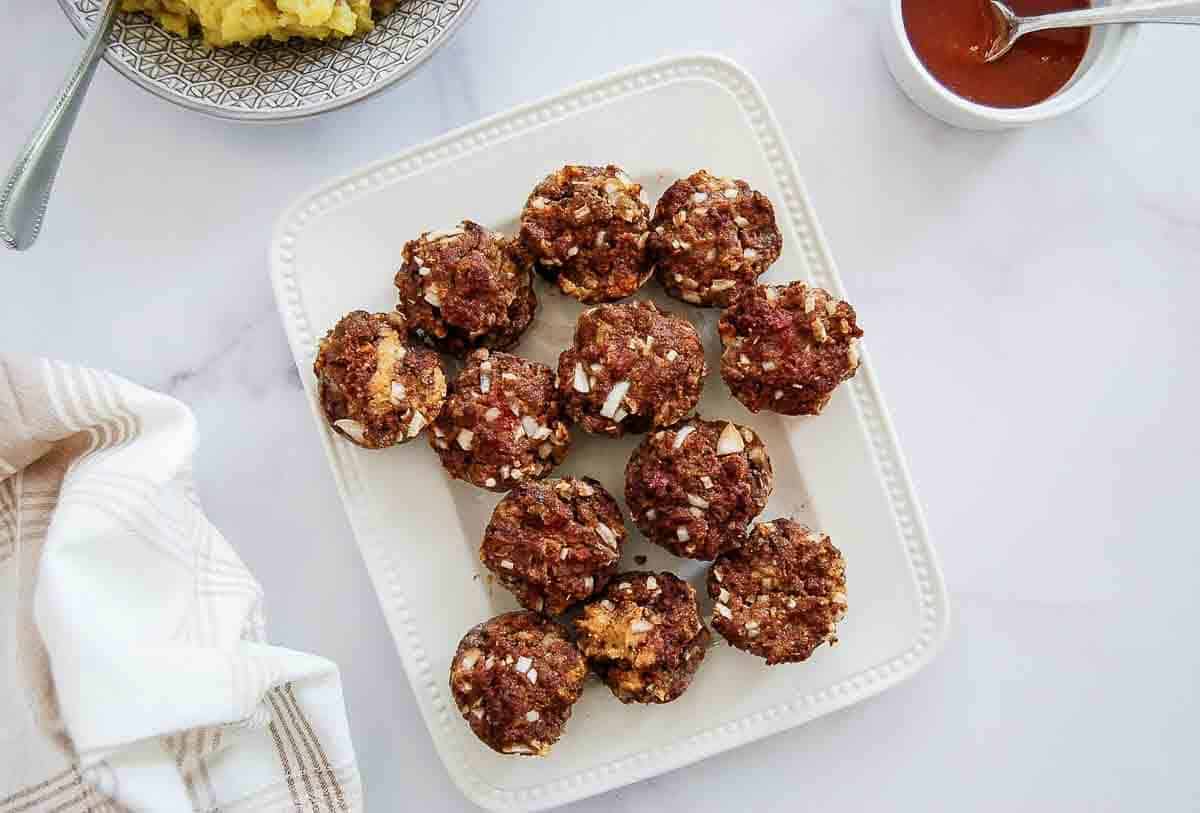 A white plate holds twelve mini meatloaf muffins. They're on a marble surface, near a bowl of mashed potatoes,.
