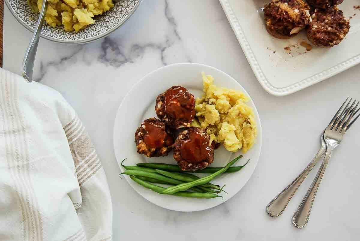A plate with three mini meatloaves topped with sauce, mashed potatoes, and green beans. Nearby are a bowl of mashed potatoes, a dish with more mini meatloaves, two forks, and a striped towel on a marble surface.