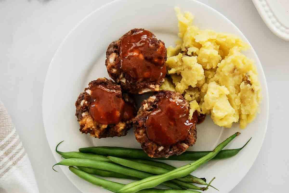 A white plate with three mini meatloafs topped with sauce, a serving of mashed potatoes, and a handful of green beans.