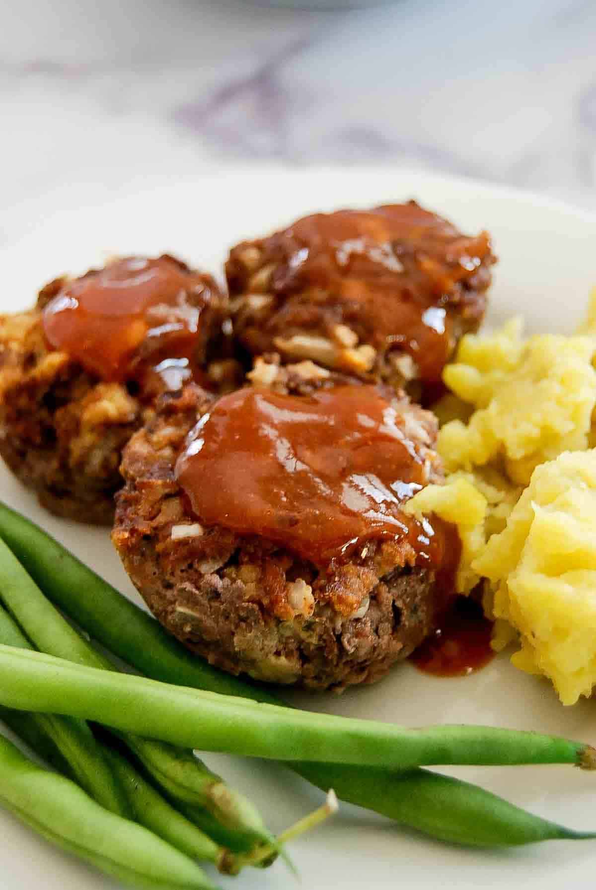 Three mini meatloaves topped with barbecue sauce are served on a white plate with a side of mashed potatoes and green beans.