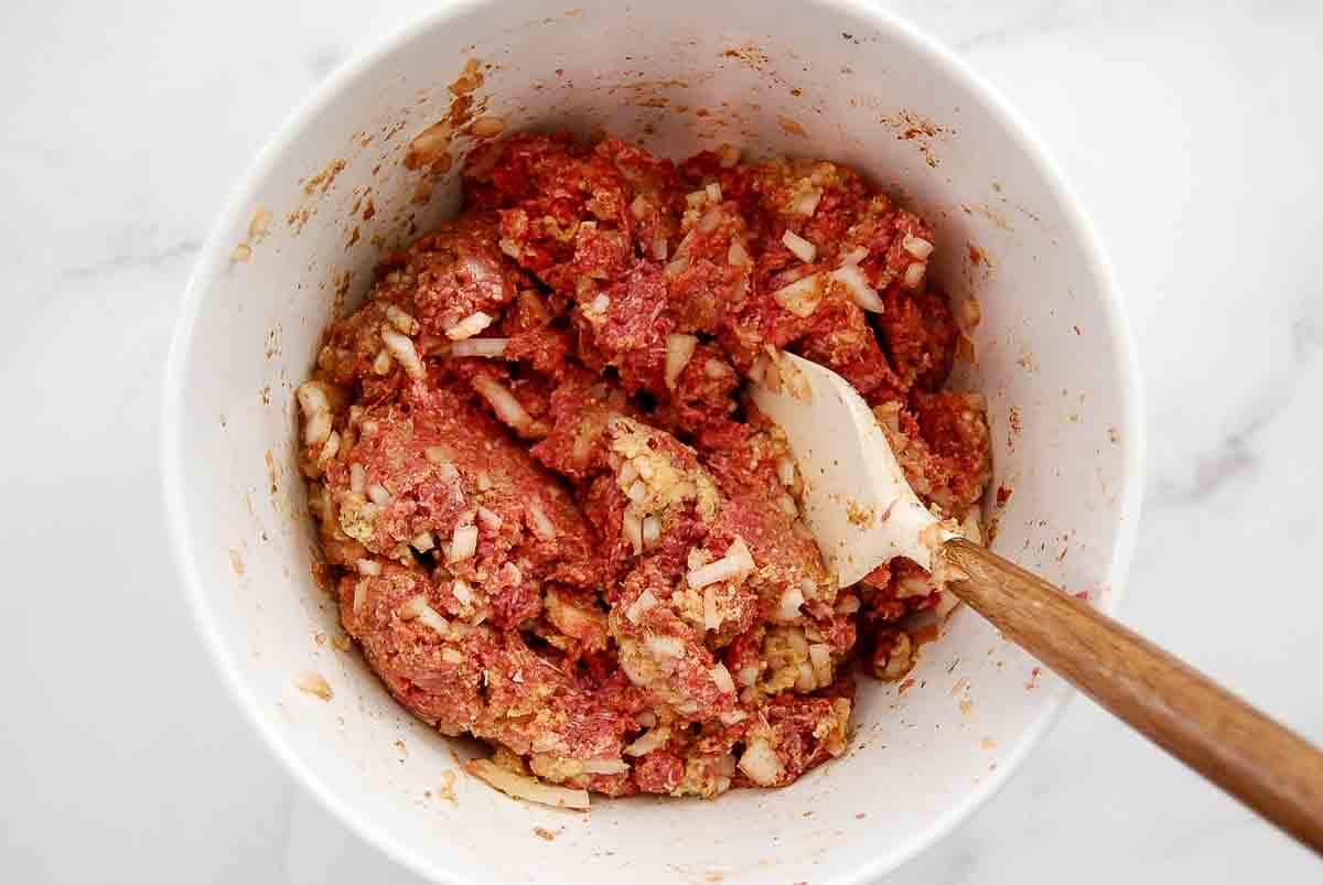 A white mixing bowl filled with raw ground meat, chopped onions, and seasonings being mixed with a wooden spatula on a light countertop.