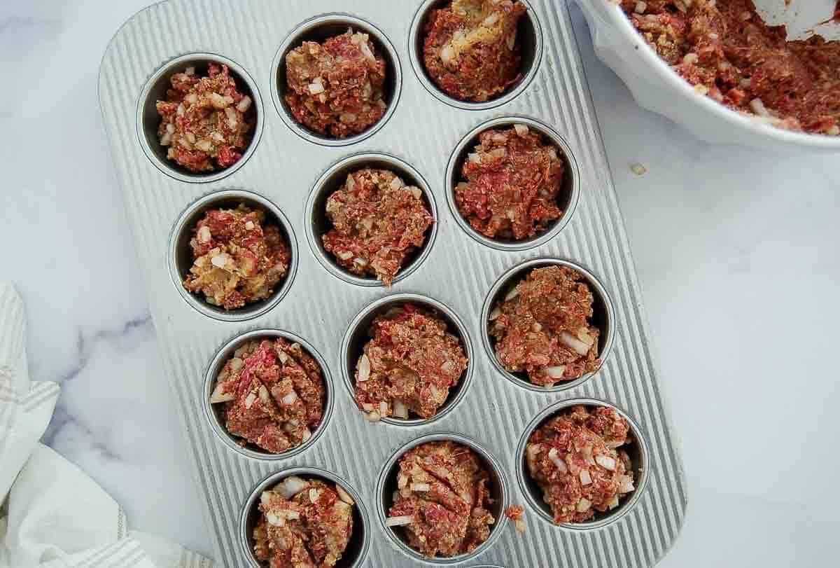 A muffin tin filled with portions of uncooked meatloaf mixture, containing visible chopped onions, sits on a white surface next to a mixing bowl with more of the same mixture.