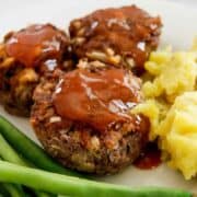 Three mini meatloaves topped with ketchup glaze are served on a plate with mashed potatoes and green beans.