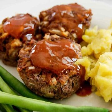 Three mini meatloaves topped with ketchup glaze are served on a plate with mashed potatoes and green beans.