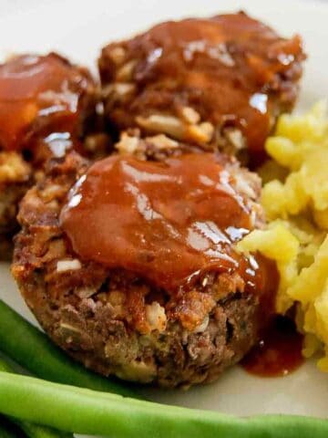 Three mini meatloaves topped with ketchup glaze are served on a plate with mashed potatoes and green beans.