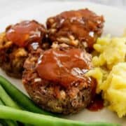 Three mini meatloaves topped with sauce are served on a plate with a side of mashed potatoes and fresh green beans.