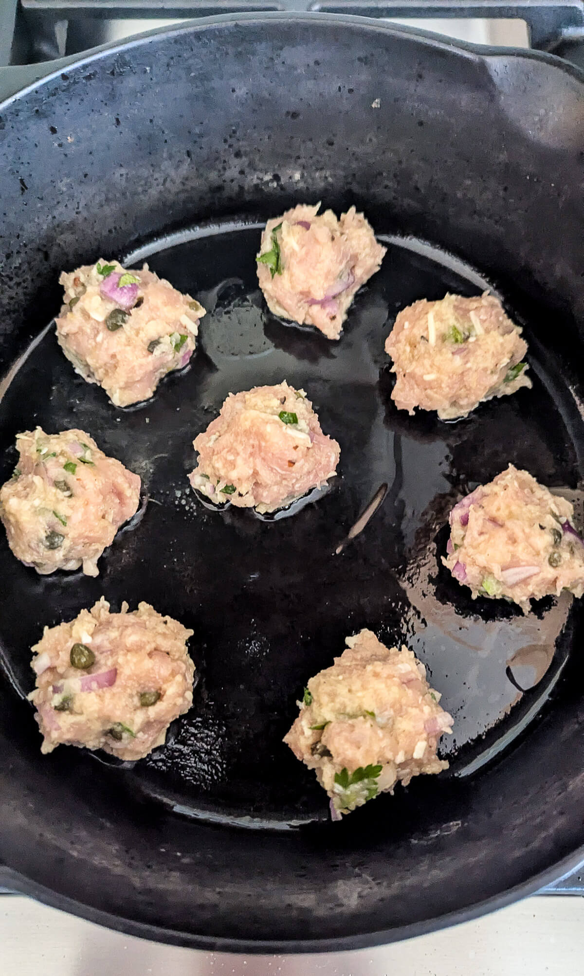 Eight raw chicken piccata meatballs with visible herbs and chopped onions are arranged in a black cast iron skillet, with a little oil, ready to be cooked.