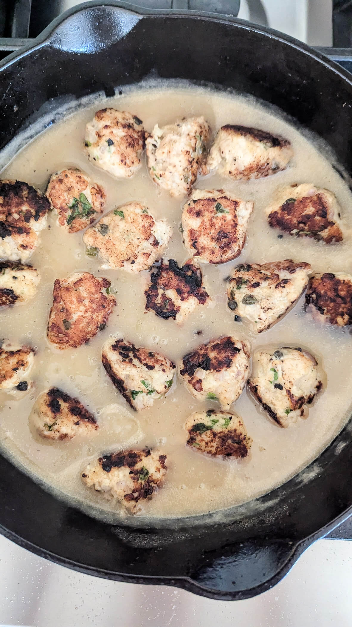 Chicken piccata meatballs simmering in a light lemon caper sauce inside a black cast iron skillet, with some meatballs browned and garnished with herbs.