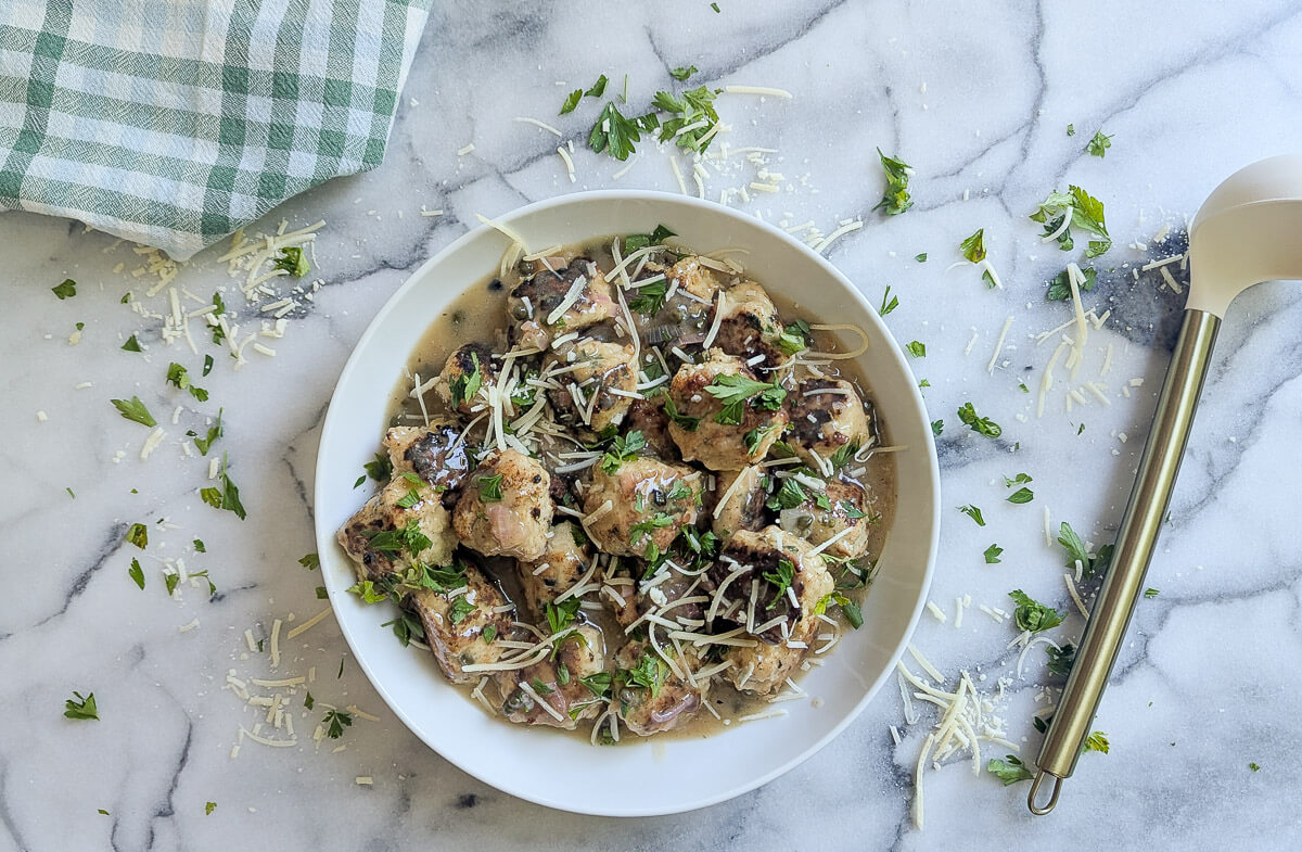 A white plate filled with chicken piccata meatballs in a creamy sauce, garnished with chopped parsley and grated cheese, sits on a marble surface.