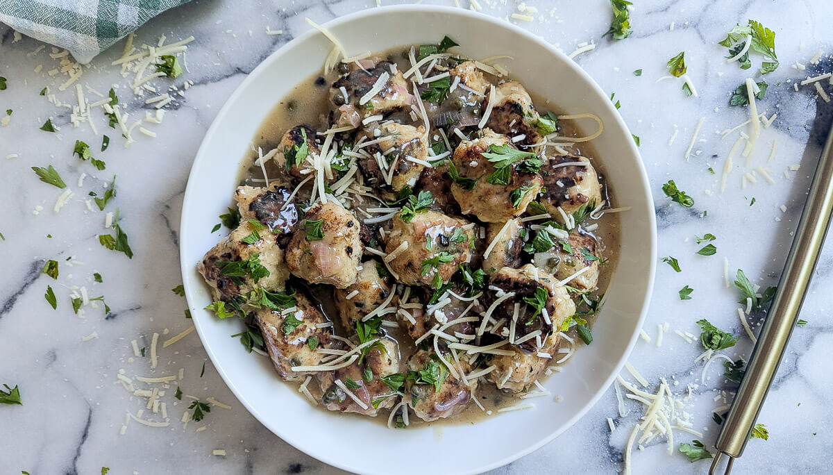 A white bowl filled with chicken piccata meatballs topped with shredded cheese and chopped parsley sits on a marble surface, with cheese and herbs scattered around.