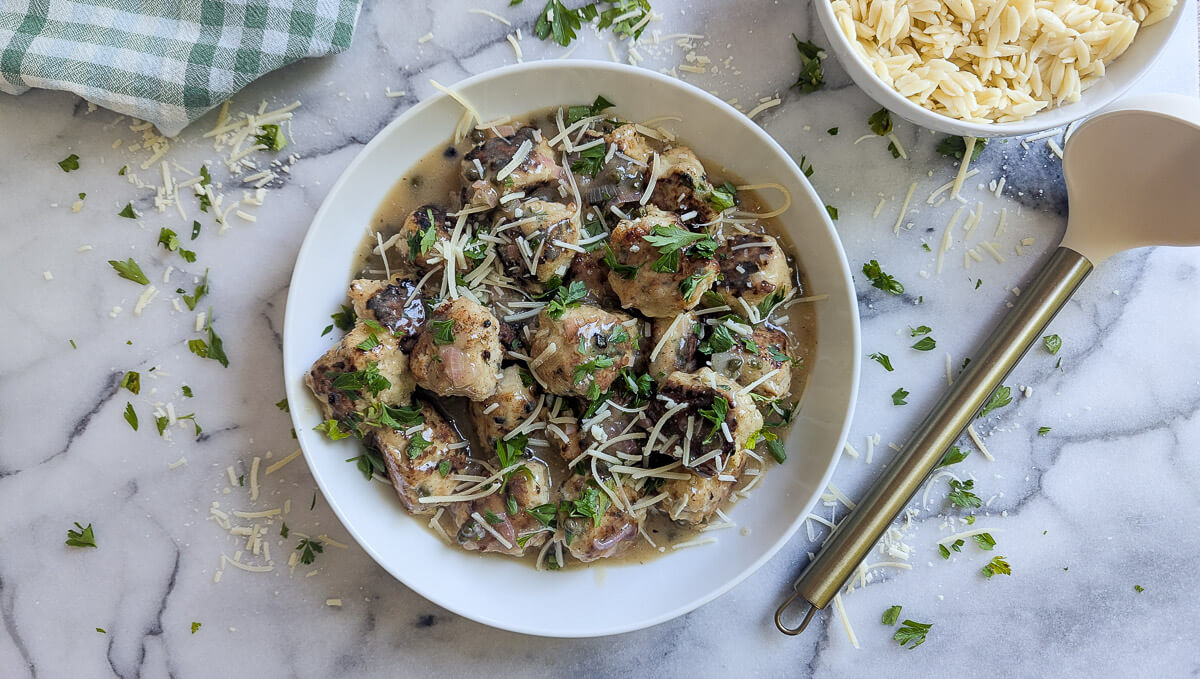 A white plate of cooked chicken piccata meatballs topped with grated cheese and chopped parsley, set on a marble surface with scattered cheese, parsley, a checkered cloth, a spoon, and a bowl of orzo nearby.