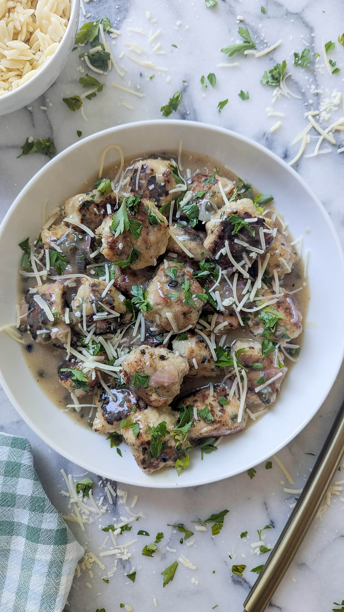 A white bowl filled with chicken piccata meatballs in lemon caper sauce, garnished with shredded cheese and fresh parsley, placed on a marble surface with scattered cheese and herbs, next to a small bowl of pasta.