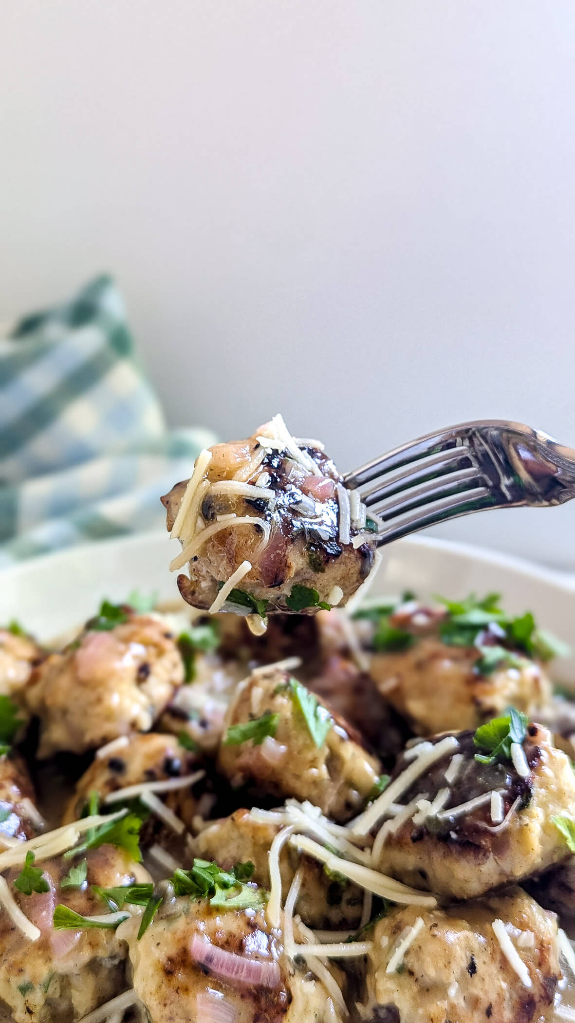 A fork holds a bite-sized chicken piccata meatball topped with shredded cheese and herbs above a plate of similar chicken meatballs, with more cheese and parsley sprinkled on top. A blurred checkered cloth is in the background.