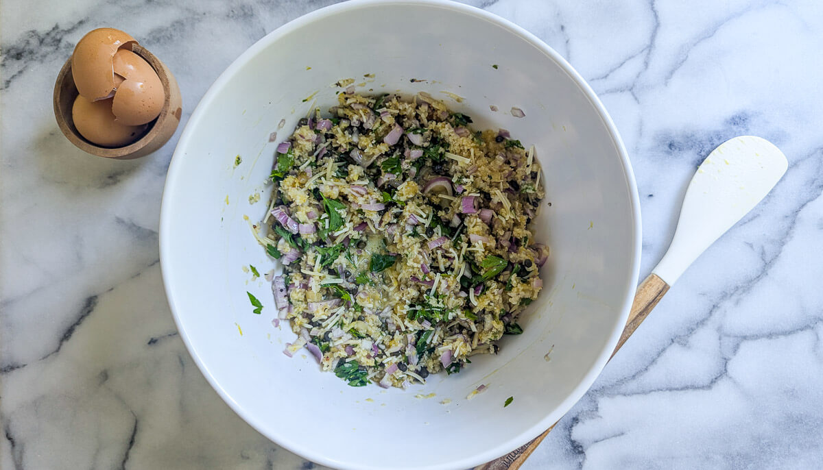 A large white bowl on a marble surface contains a mixture of chopped herbs, breadcrumbs, egg, capers, shallot, garlic, and cheese.