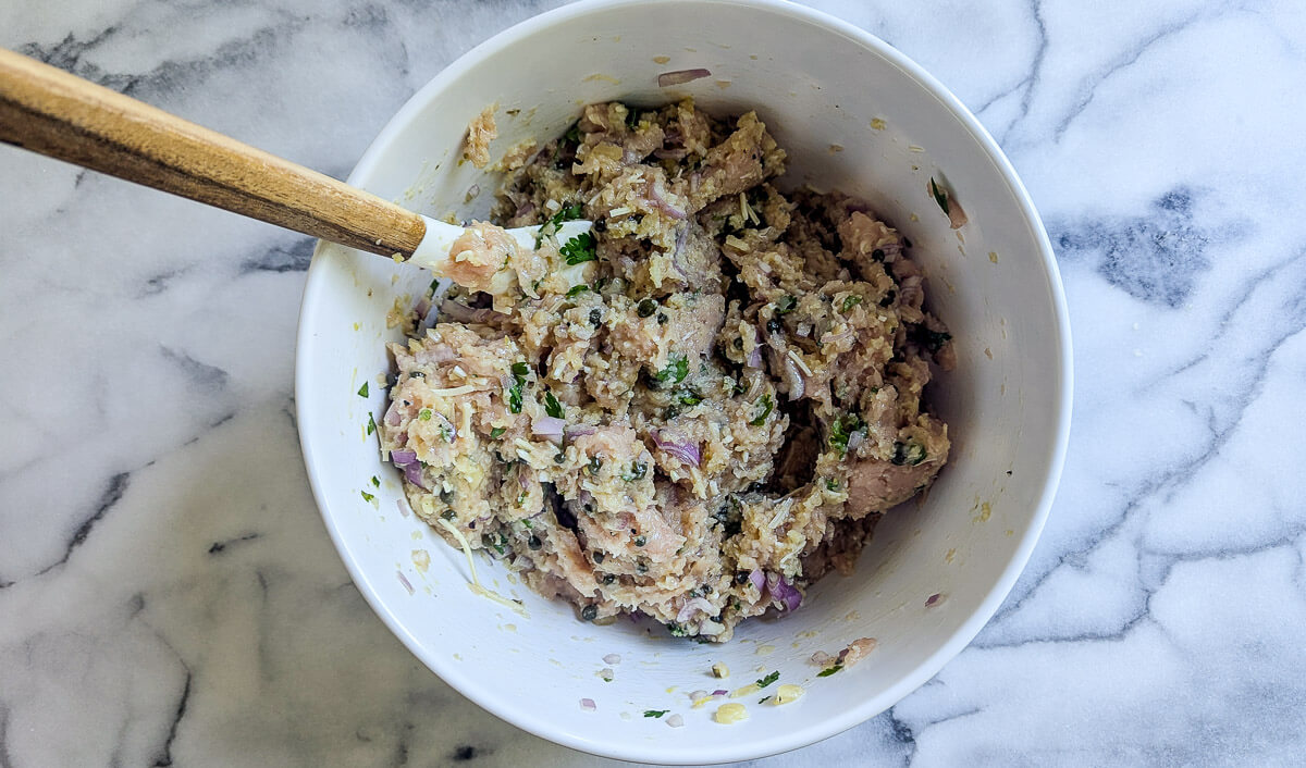 A white bowl containing a mixture of ground chicken, chopped herbs, onions, and seasonings sits on a marble countertop. A wooden-handled spatula rests inside the bowl, partially covered with the mixture.