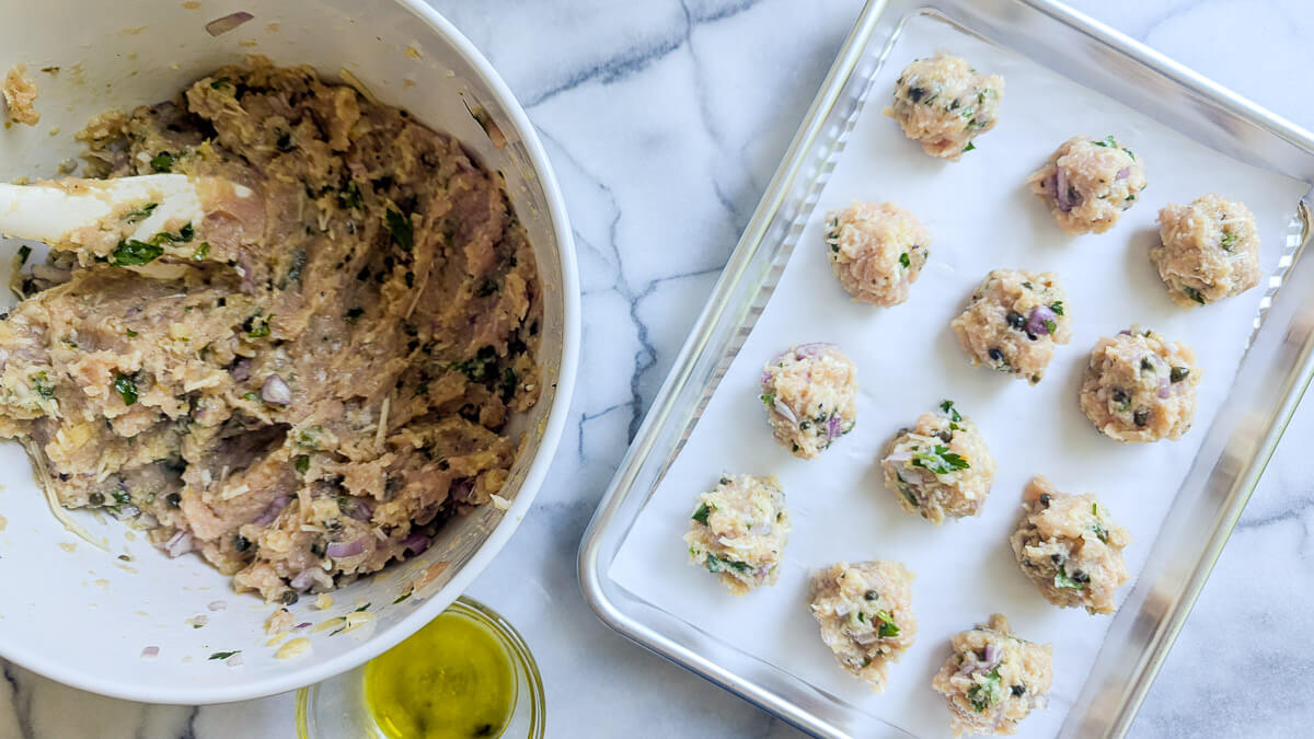 A bowl of raw chicken meatball mixture with herbs next to a tray of unbaked meatballs, ready for the oven. A small bowl of olive oil sits nearby on a marble countertop.