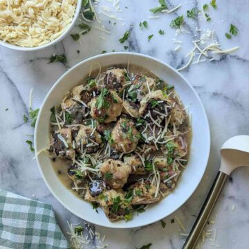 A white bowl filled with chicken piccata meatballs in lemon caper sauce, topped with shredded cheese and chopped parsley, sits on a marble surface beside a bowl of orzo, grated cheese, and a green checked napkin.
