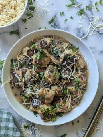 A white bowl filled with chicken piccata meatballs in lemon caper sauce, topped with shredded cheese and chopped parsley, sits on a marble surface beside a bowl of orzo, grated cheese, and a green checked napkin.