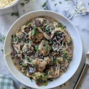 A white plate of chicken piccata meatballs in lemon caper sauce, garnished with chopped parsley and shredded cheese, on a marble surface with a bowl of orzo and a checkered napkin nearby.