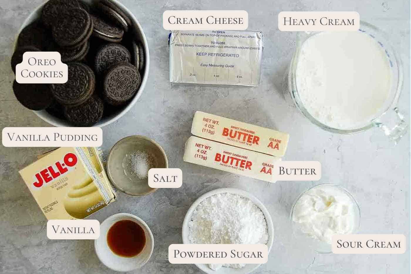 Ingredients for a dessert are arranged on a light surface: Oreo cookies, vanilla pudding mix, vanilla extract, cream cheese, heavy cream, sour cream, butter, powdered sugar, and salt, each labeled with text.