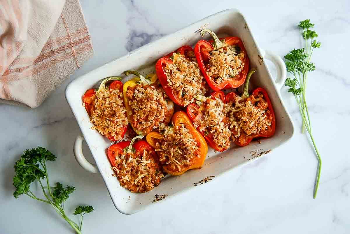 A white baking dish filled with halved red bell peppers stuffed with ground turkey, quinoa, marinara, and topped with a breadcrumb mixture, placed on a marble countertop with parsley sprigs and a striped cloth nearby.