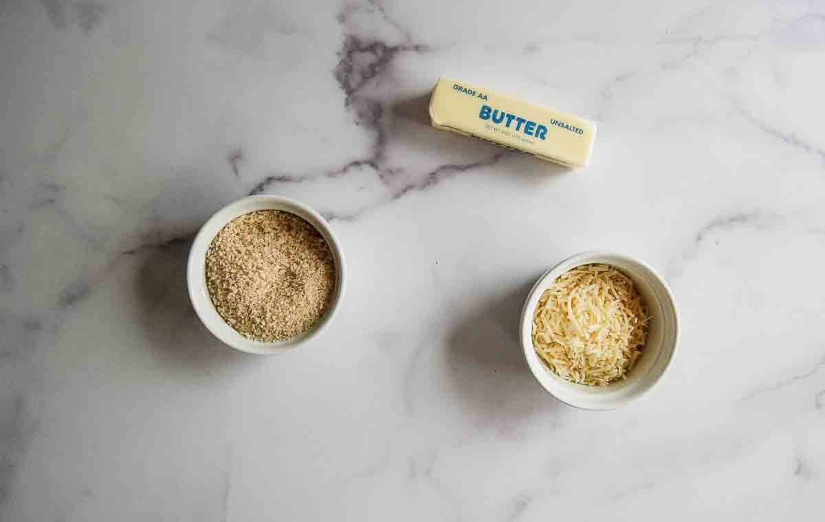 A stick of butter, a bowl of breadcrumbs, and a bowl of shredded cheese are arranged on a white marble surface.