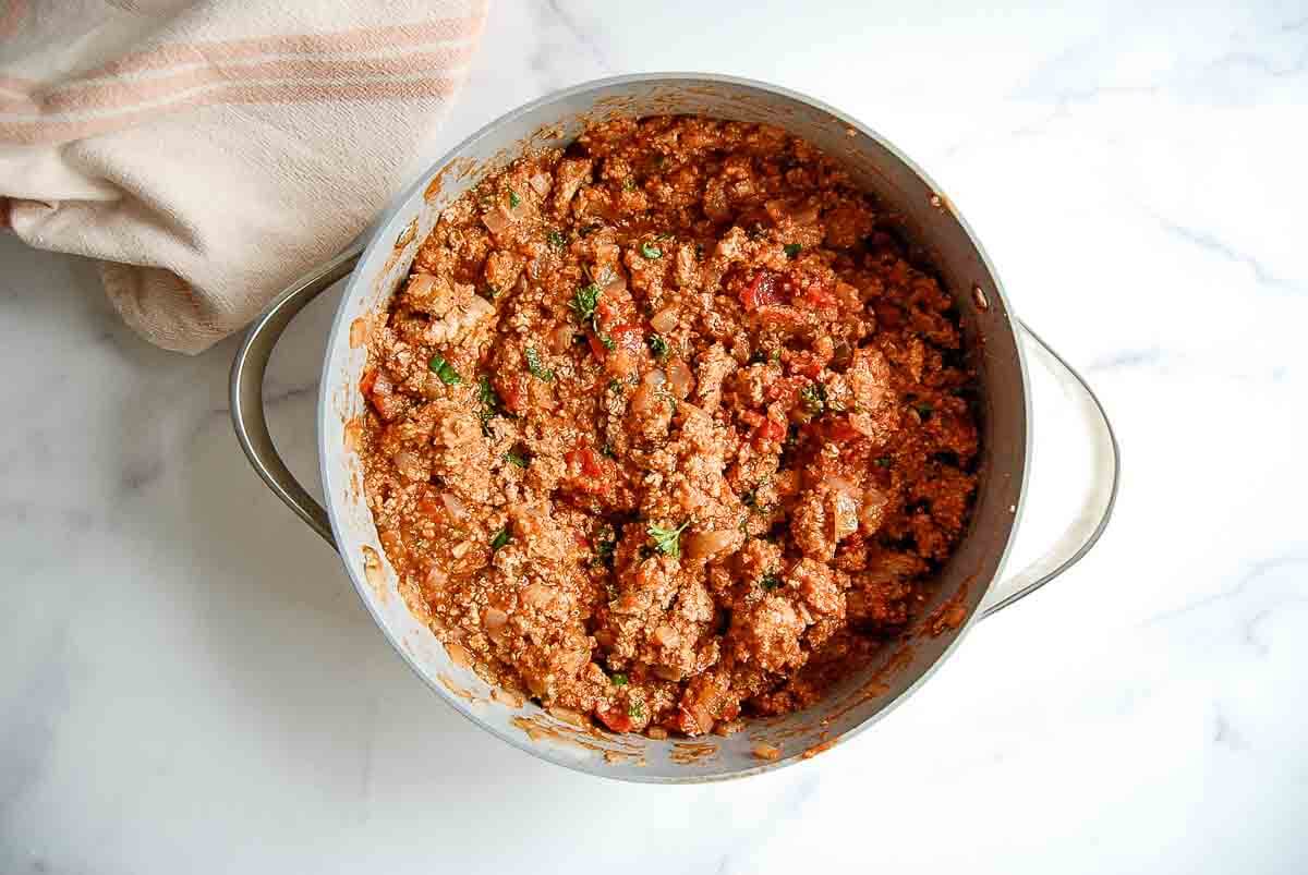 A large pot filled with cooked quinoa and ground turkey, mixed with tomato sauce, herbs, and vegetables sits on a white marble surface next to a folded beige and pink striped towel.