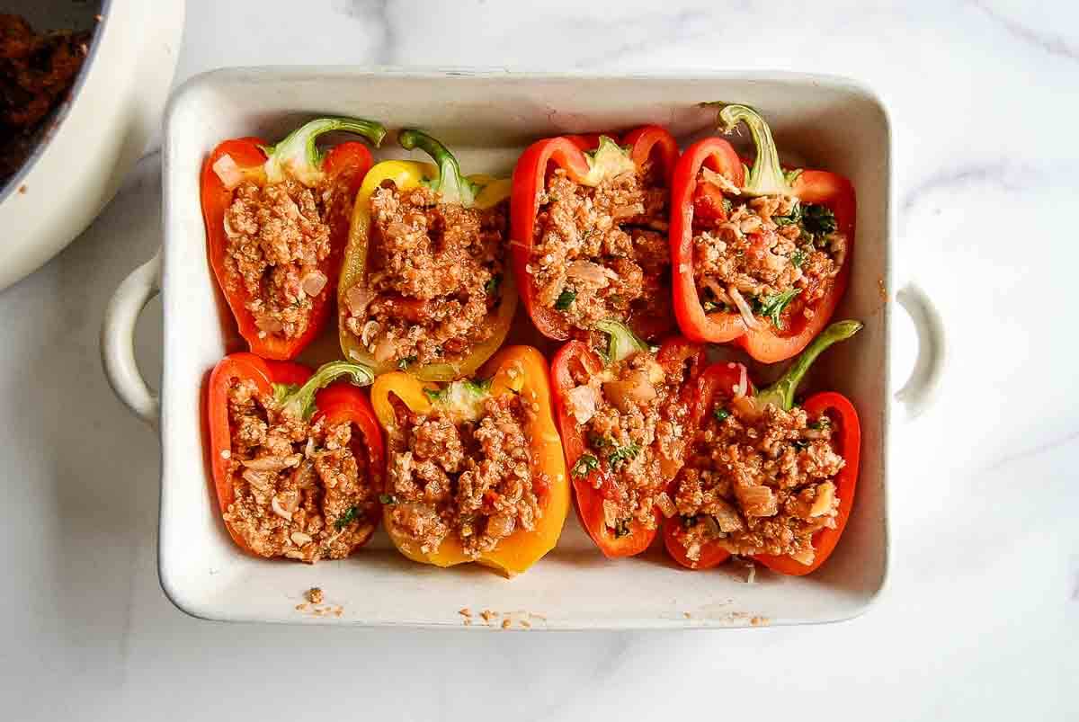 A white baking dish filled with halved red and yellow bell peppers, each stuffed with a seasoned ground turkey mixture, on a white marble surface.