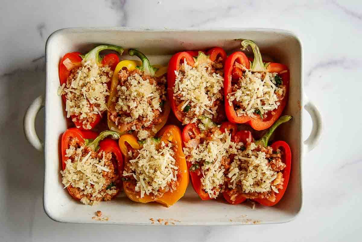 A baking dish filled with halved red and yellow bell peppers, stuffed with a mixture of ground turkey, marinara sauce, vegetables, and topped with shredded cheese and breadcrumbs, ready to be baked.