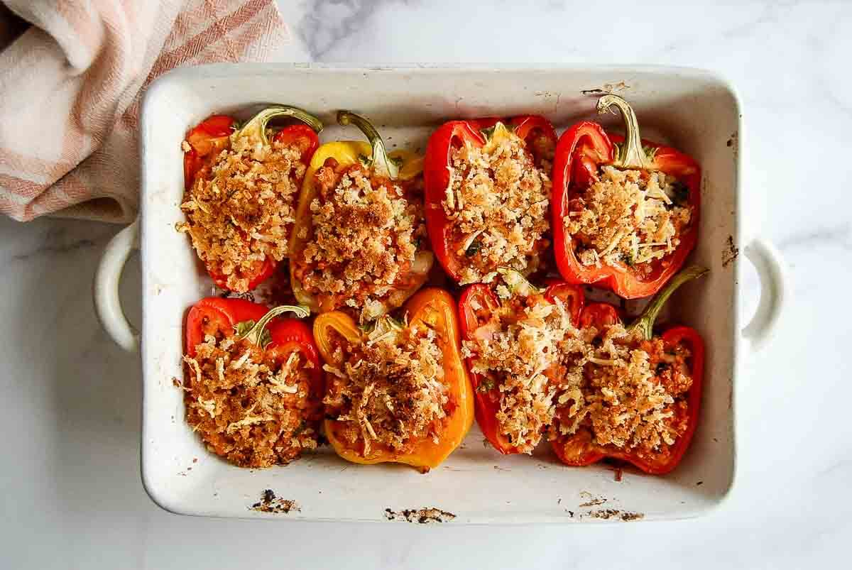 Halved red and yellow bell peppers stuffed with a ground turkey mixture, topped with a breadcrumb and grated cheese mixture, arranged in a white baking dish on a marble surface.