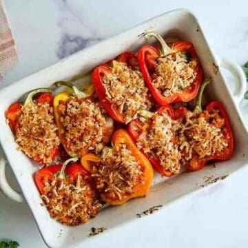 A white baking dish holds eight stuffed bell pepper halves, filled with a breadcrumb and cheese topping. The peppers are red and yellow, arranged neatly and baked until golden brown on top, set on a marble countertop.