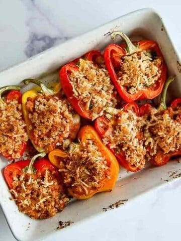 A white baking dish holds eight stuffed bell pepper halves, filled with a breadcrumb and cheese topping. The peppers are red and yellow, arranged neatly and baked until golden brown on top, set on a marble countertop.
