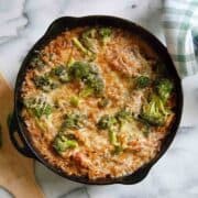 A cast iron skillet filled with a lemon chicken orzo casserole featuring melted cheese, broccoli florets, and orzo pasta, sitting on a marble surface next to a green and white checkered cloth.
