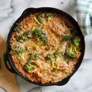 A cast iron skillet filled with a lemon chicken orzo casserole featuring melted cheese, broccoli florets, and orzo pasta, sitting on a marble surface next to a green and white checkered cloth.