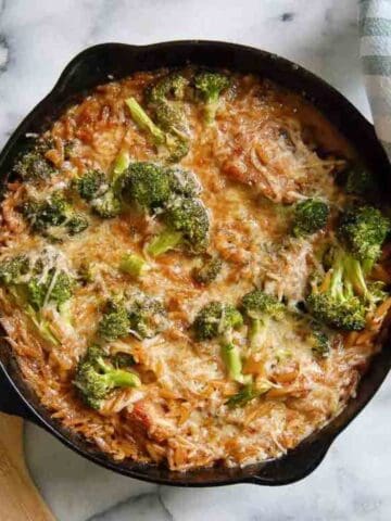 A cast iron skillet filled with a lemon chicken orzo casserole featuring melted cheese, broccoli florets, and orzo pasta, sitting on a marble surface next to a green and white checkered cloth.