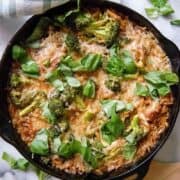 A skillet lemon chicken orzo casserole topped with melted cheese, broccoli florets, and fresh basil leaves, sits on a marble surface with a green checkered cloth and chopped basil nearby.