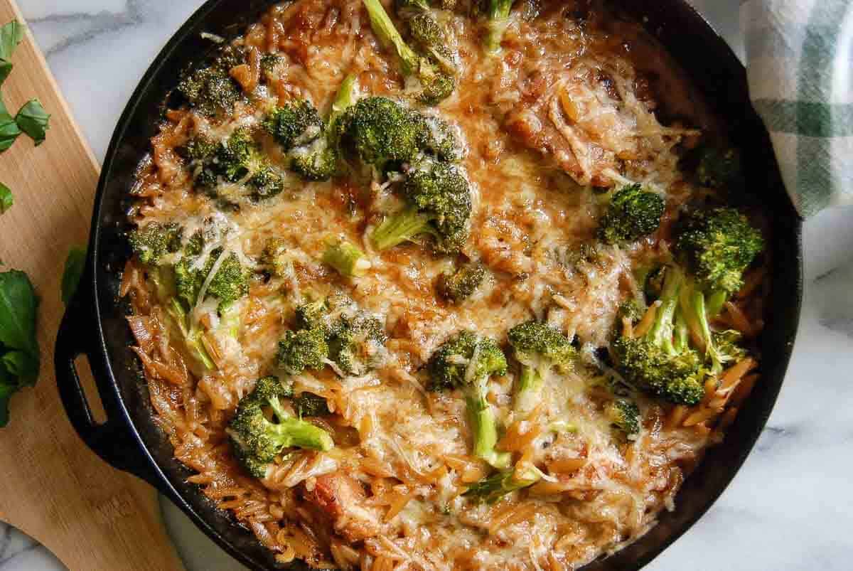 A skillet filled with lemon chicken orzo, topped with melted cheese and broccoli florets, sits on a marble surface next to a wooden board with fresh herbs and a white and green kitchen towel.