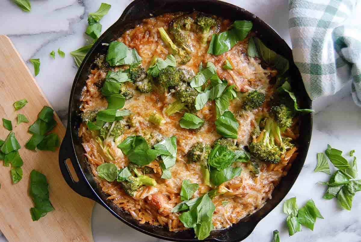 A cast iron skillet filled with baked orzo, chicken and lemon, topped with melted cheese, broccoli florets, and fresh basil leaves, sits on a marble surface next to a wooden board with more basil and a green-checked cloth.