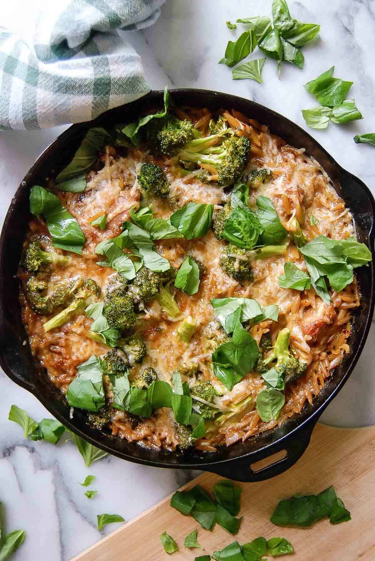 A cast iron skillet filled with lemon chicken and orzo topped with broccoli, melted cheese, and fresh basil leaves, sitting on a marble countertop with a green plaid towel and scattered basil nearby.