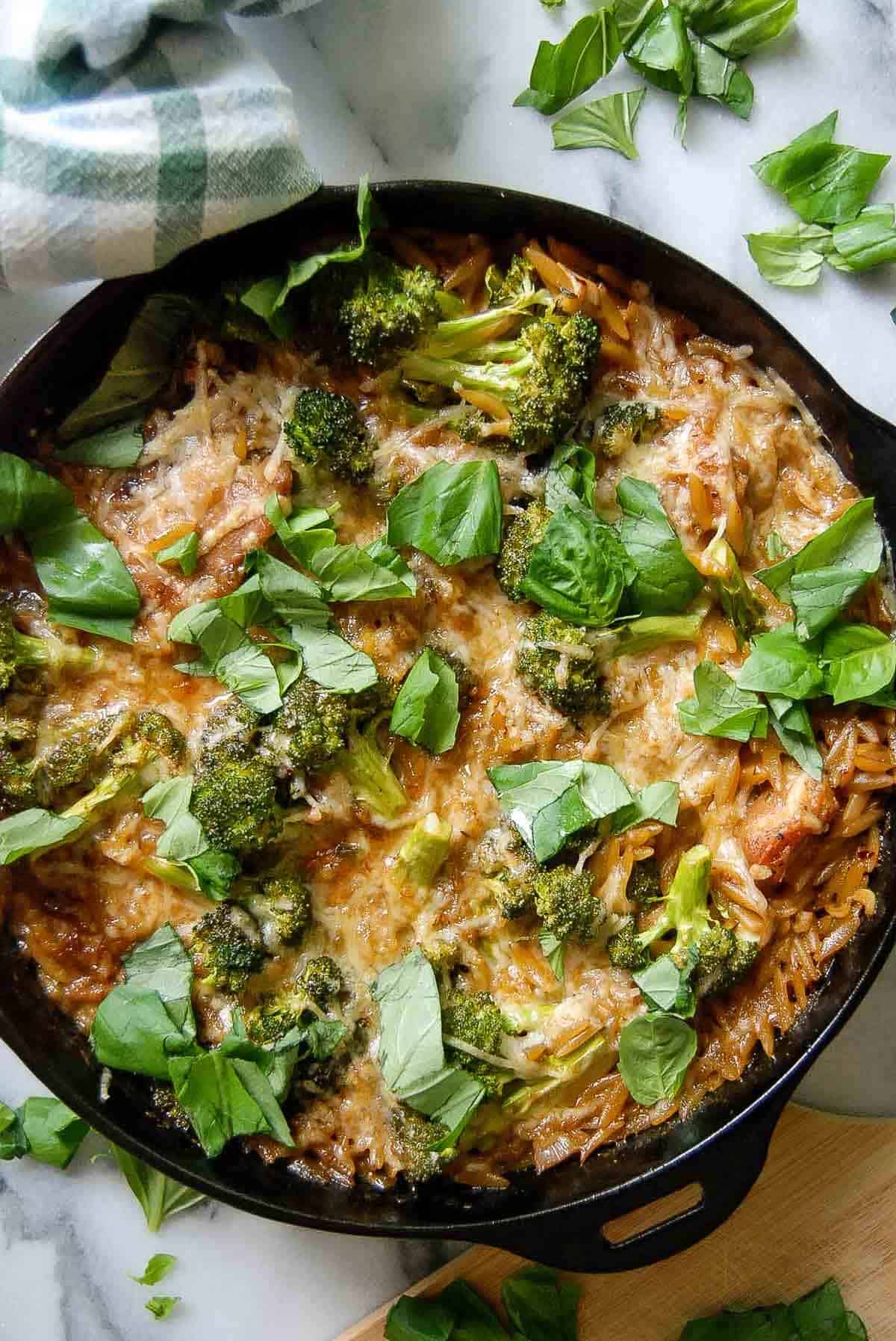 A cast iron skillet filled with chicken and orzo topped with melted cheese, broccoli florets, and fresh basil leaves, sitting on a marble surface with a checkered cloth nearby.